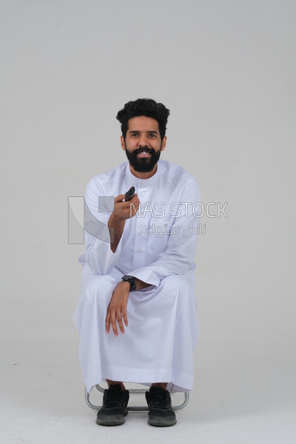 Saudi man wearing a Saudi dress, sitting on a chair, holding the remote control in his hand, spending good time, watching TV, happy facial gestures, spending fun times, entertainment, movies and cin