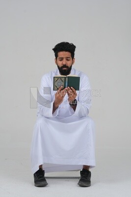 Saudi man wearing a white dress sitting on a chair, reading Quran, Islam and worship, Saudi model, white background