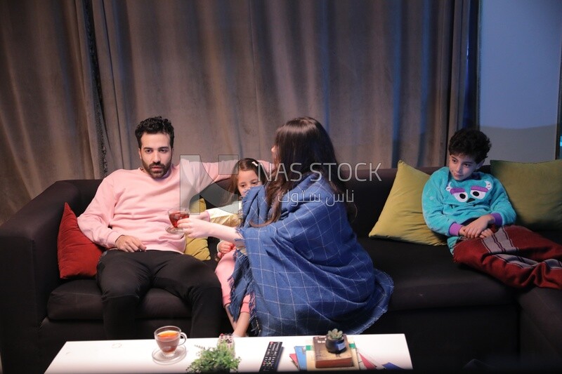 a family of four watching television and the mother gives the father a cup of tea