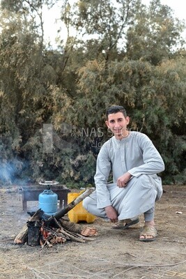 صورة عن قرب لشاب يرتدى جلباب رمادى يشعل حطب فى خلفية لاشجار