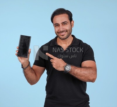 Photo Of a Handsome man Standing and holding his mobile pointing on it, Smiling Man, Wearing Casual Clothes Posing In a Light Room Interior, Smiling At the Camera