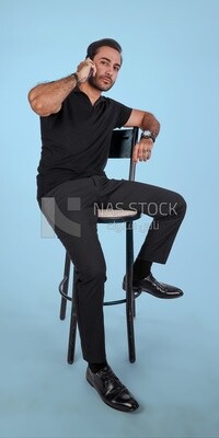 Photo of a handsome man sitting and talking to the mobile phone, man smiling, dressed in casual clothes in light room interior, smiling at camera