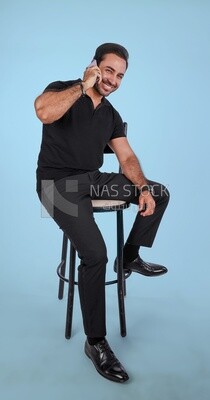 Photo of a handsome man sitting and talking to the mobile phone, man smiling, dressed in casual clothes in light room interior, smiling at camera