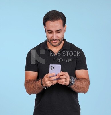 صورة لرجل وسيم يقف ويمسك هاتفه المحمول ، رجل مبتسم ، يرتدي ملابس غير رسمية ، يقف في داخل غرفة خفيفة ، ويبتسم للكاميرا