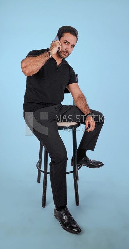 Photo of a handsome man sitting and talking to the mobile phone, man smiling, dressed in casual clothes in light room interior, smiling at camera