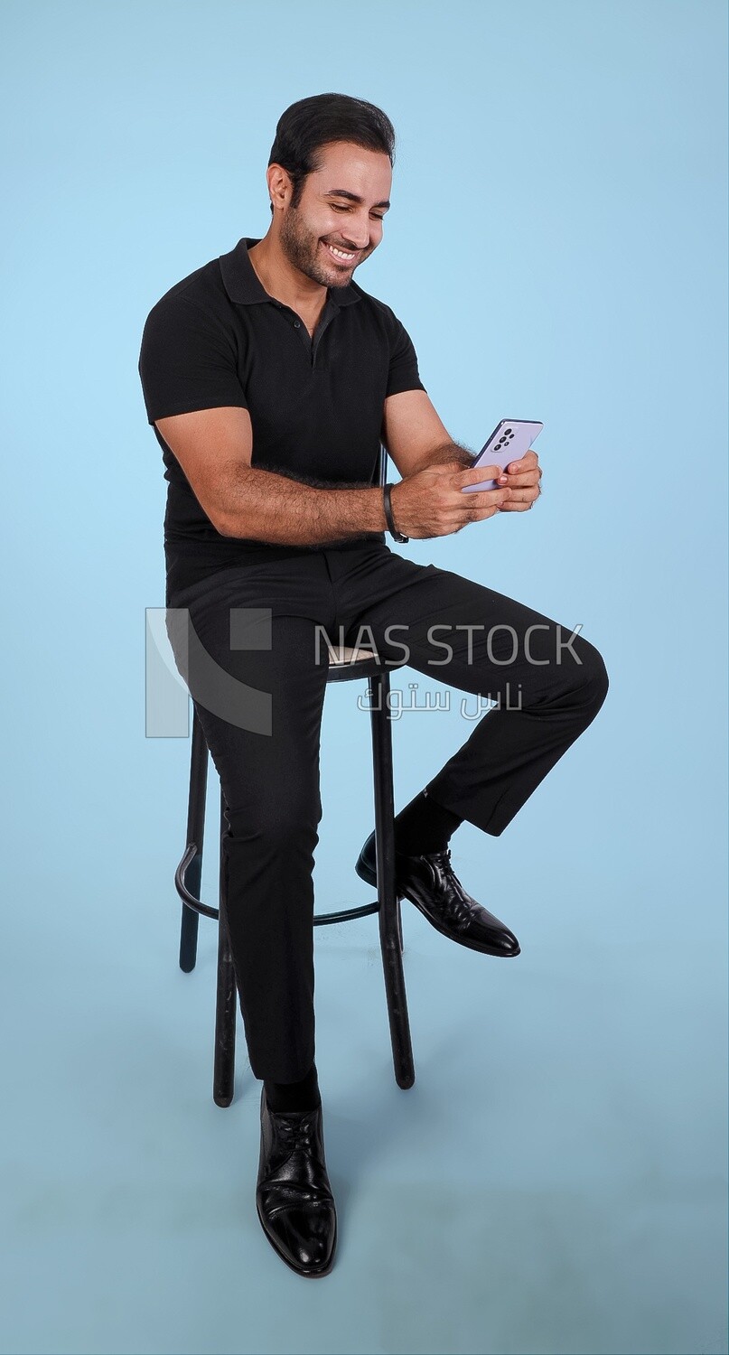 Photo Of a Handsome man sitting on the chair and holding his mobile, Smiling Man, Wearing Casual Clothes Posing In a Light Room Interior, Smiling At the Camera