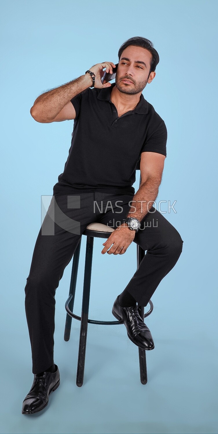 Photo of a handsome man sitting and talking to the mobile phone, man smiling, dressed in casual clothes in light room interior, smiling at camera
