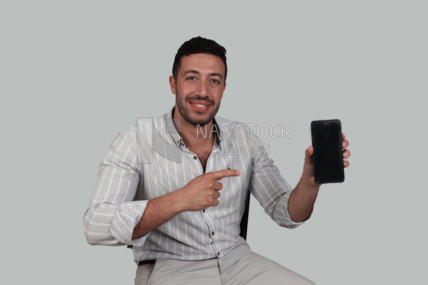 Photo Of a Handsome man sitting on a chair holding his mobile, Smiling Man, Wearing Casual Clothes Posing In a Light Room Interior, Smiling At the Camera, looking away