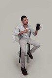 Photo Of a Handsome man sitting on a chair holding his mobile, Smiling Man, Wearing Casual Clothes Posing In a Light Room Interior, Smiling At the Camera, looking away