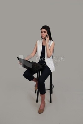 Photo of a businesswoman with formal wear sitting on a chair working on a laptop while talking on the phone, business development and partnerships, business meeting, Model