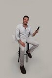 Photo Of a Handsome man sitting on a chair checking his mobile, Smiling Man, Wearing Casual Clothes Posing In a Light Room Interior, Smiling At the Camera, looking away