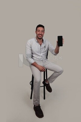 Photo Of a Handsome man sitting on a chair holding his mobile, Smiling Man, Wearing Casual Clothes Posing In a Light Room Interior, Smiling At the Camera, looking away