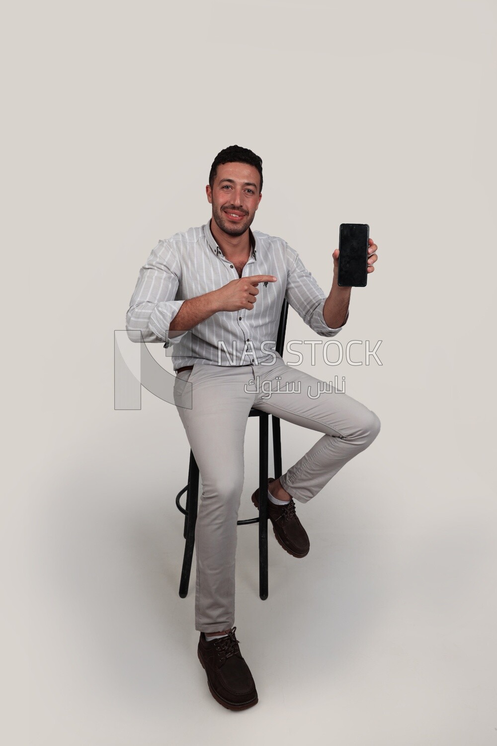 Photo Of a Handsome man sitting on a chair holding his mobile, Smiling Man, Wearing Casual Clothes Posing In a Light Room Interior, Smiling At the Camera, looking away