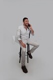 Photo Of a Handsome man sitting on a chair talking on his mobile, Smiling Man, Wearing Casual Clothes Posing In a Light Room Interior, Smiling At the Camera, looking away