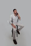 Photo Of a Handsome man sitting on a chair talking on his mobile, Smiling Man, Wearing Casual Clothes Posing In a Light Room Interior, Smiling At the Camera, looking away