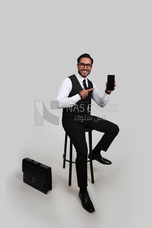 Photo of a businessman in formal suit sitting on a chair and holding a mobile phone pointing at it, business development and partnerships, business meeting, Model