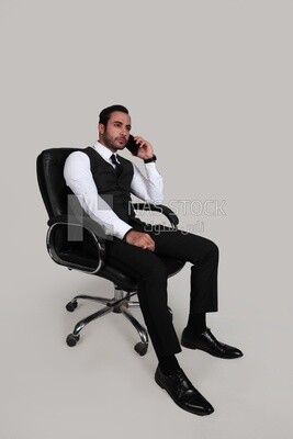 Photo of a businessman with a formal suit sitting on a chair and talking in his mobile phone, business development and partnerships, business meeting, Model