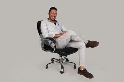 Photo Of a Handsome man sitting on a chair putting a leg on a leg, Smiling Man, Wearing Casual Clothes Posing In a Light Room Interior, Smiling At the Camera, looking away