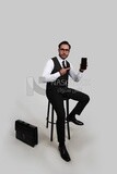 Photo of a businessman in formal suit sitting on a chair and holding a mobile phone pointing at it, business development and partnerships, business meeting, Model