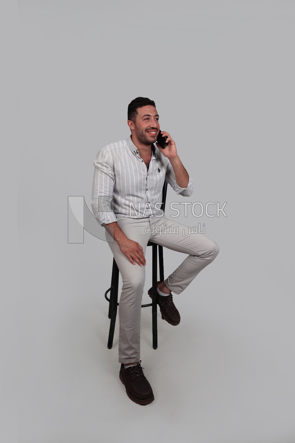Photo Of a Handsome man sitting on a chair and talking on his phone, Smiling Man, Wearing Casual Clothes Posing In a Light Room Interior, Smiling At the Camera, looking away