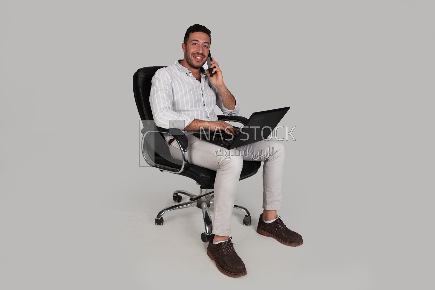 Photo of a businessman with formal wear sitting on a chair working on a laptop while talking on the phone, business development and partnerships, business meeting, Model