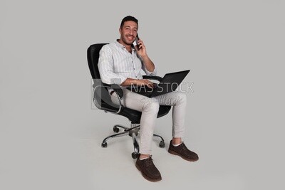 Photo of a businessman with formal wear sitting on a chair working on a laptop while talking on the phone, business development and partnerships, business meeting, Model