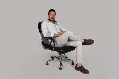 Photo of a handsome man sitting on a chair putting a leg on a leg, Smiling Man, Wearing Casual Clothes Posing In a Light Room Interior, Smiling At the Camera, looking away