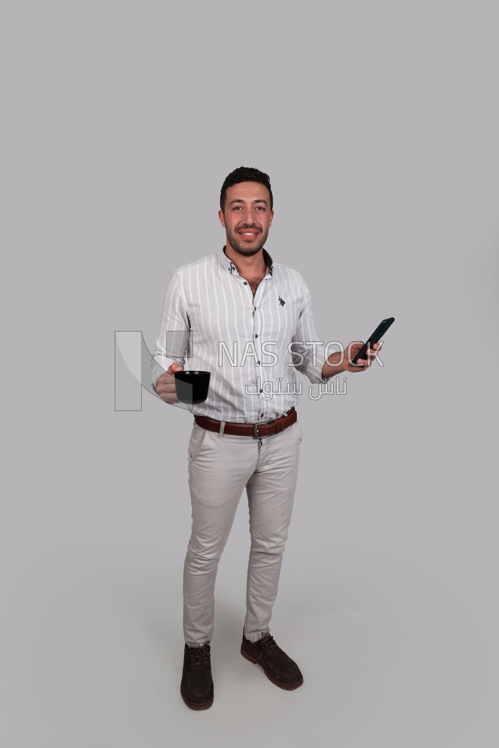 Photo Of a Handsome man holding his mobile in one hand and a cup in the other, Smiling Man, Wearing Casual Clothes Posing In a Light Room Interior, Smiling At the Camera, looking away