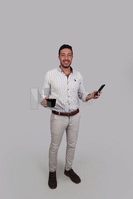 صورة لرجل وسيم يحمل هاتفه المحمول في إحدى يديه وفنجانًا في اليد الأخرى ، رجل مبتسم ، يرتدي ملابس غير رسمية في غرفة مضيئة من الداخل ، يبتسم للكاميرا ، وينظر بعيدًا