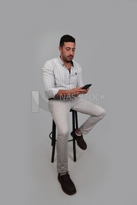 صورة لرجل وسيم يجلس على كرسي يتفحص هاتفه المحمول ، رجل يبتسم ، يرتدي ملابس غير رسمية ، يقف في داخل غرفة خفيفة ، يبتسم للكاميرا ، وينظر بعيدًا