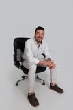 Photo Of a Handsome man sitting on a chair, Smiling Man, Wearing Casual Clothes Posing In a Light Room Interior, Smiling At the Camera, looking away