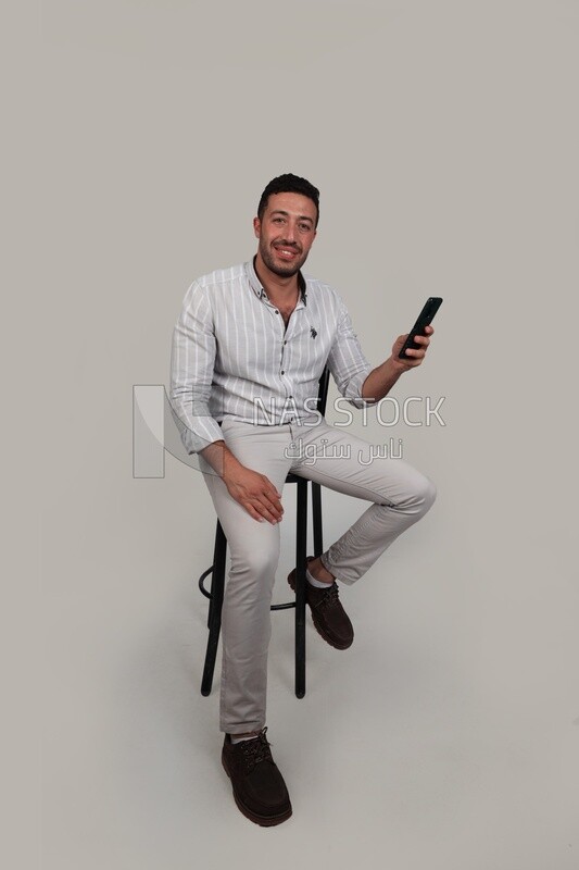 Photo Of a Handsome man sitting and checking his mobile, Smiling Man, Wearing Casual Clothes Posing In a Light Room Interior, Smiling At the Camera, looking away