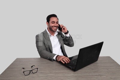 صورة لرجل أعمال يرتدي بدلة رسمية يجلس على مكتب يعمل على كمبيوتر محمول أثناء التحدث على هاتفه المحمول ، وتطوير الأعمال والشراكات ، واجتماع العمل ، موديل