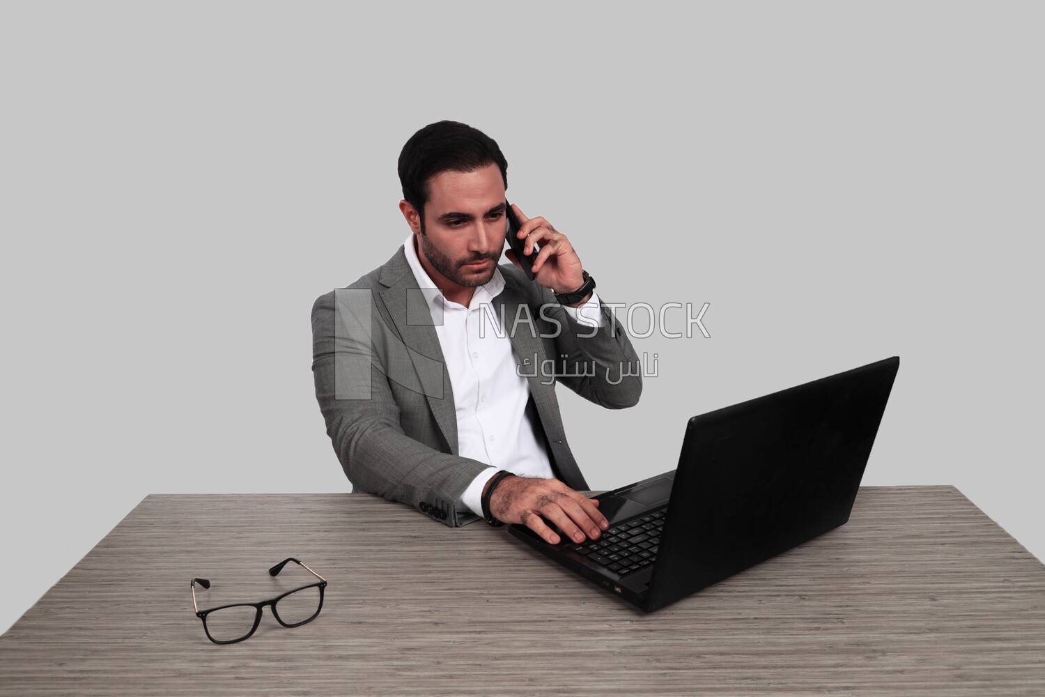 Photo of a businessman with a formal suit sitting at a desk working on a laptop while talking on the phone, business development and partnerships, business meeting, Model