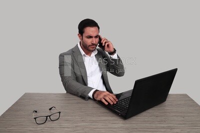 Photo of a businessman with a formal suit sitting at a desk working on a laptop while talking on his mobile, business development and partnerships, business meeting, Model