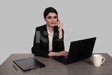 Photo of a businesswoman with a formal suit sitting at a desk working on a laptop while talking on the phone, business development and partnerships, business meeting, Model