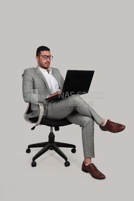 صورة لرجل أعمال يرتدي بدلة رسمية يجلس على كرسي ويضع ساقه على ساقه ويعمل على كمبيوتر محمول ، وتطوير الأعمال والشراكات ، واجتماع عمل ، موديل