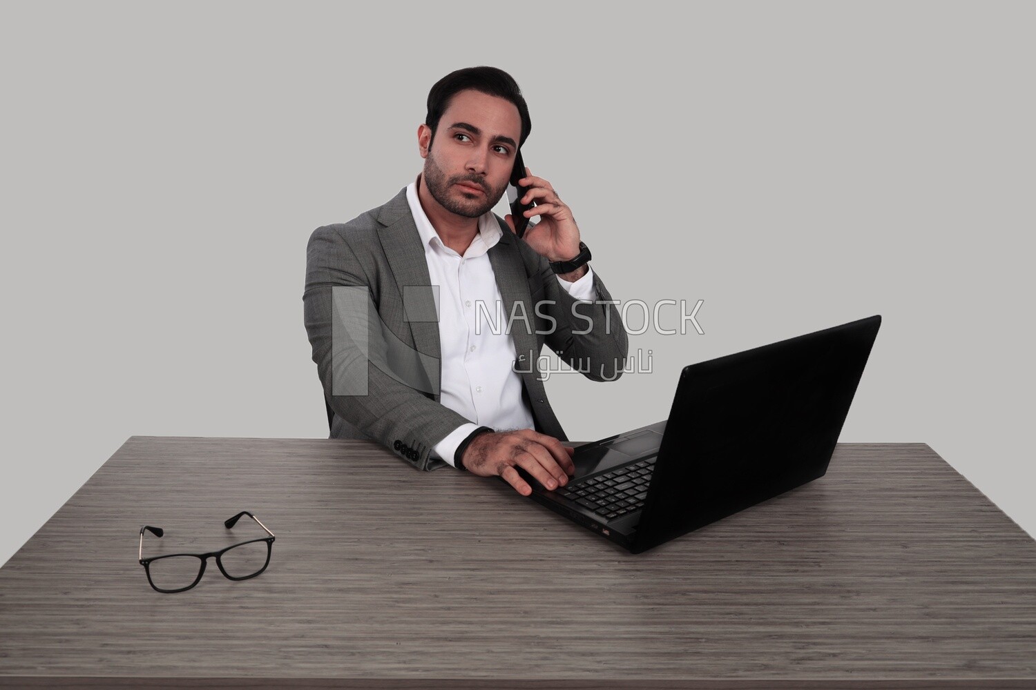 Photo of a businessman with a formal suit sitting at a desk working on a laptop while talking on his mobile, business development and partnerships, business meeting, Model