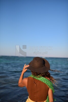 Woman wearing summer clothes and a straw hat stands in front of the sea