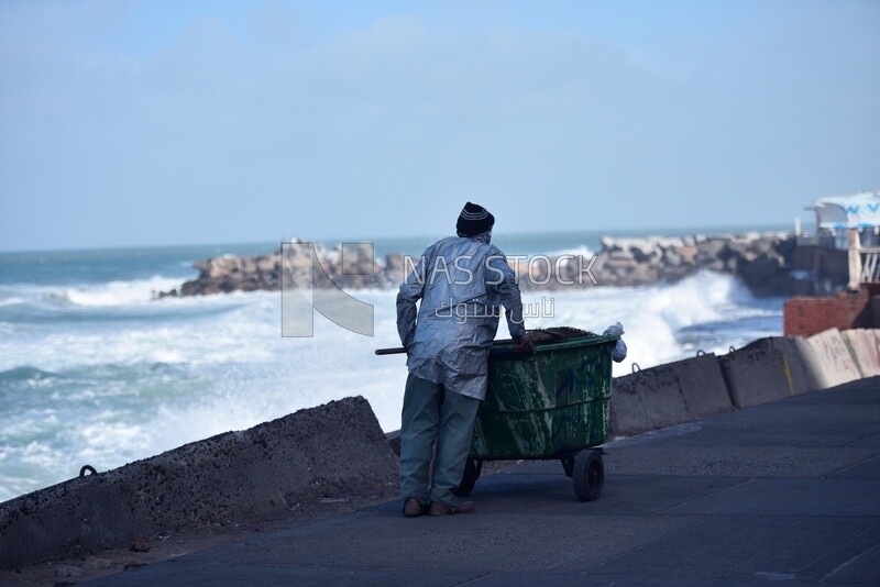 Cleaner drags a garbage can and walks on the seashore