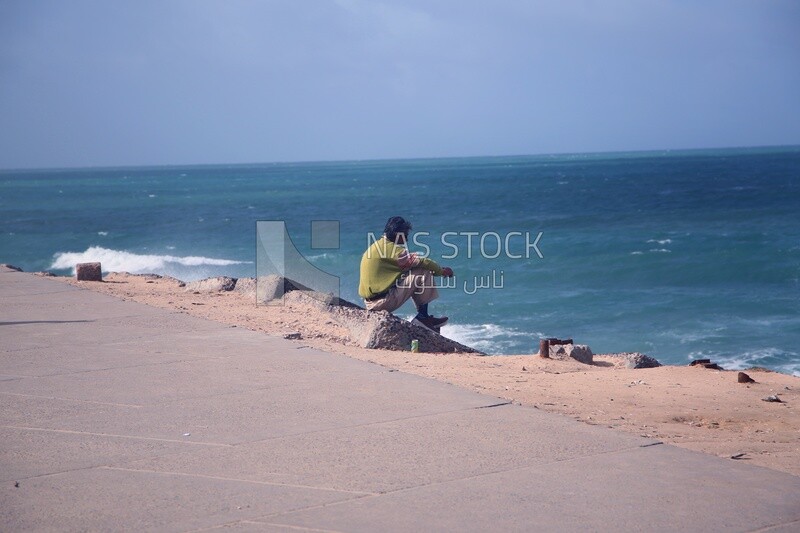 رجل بالغ يجلس على ساحل البحر يتأمل المنظر الخلاب للامواج فى البحر مع السماء , الاسكندرية , مصر