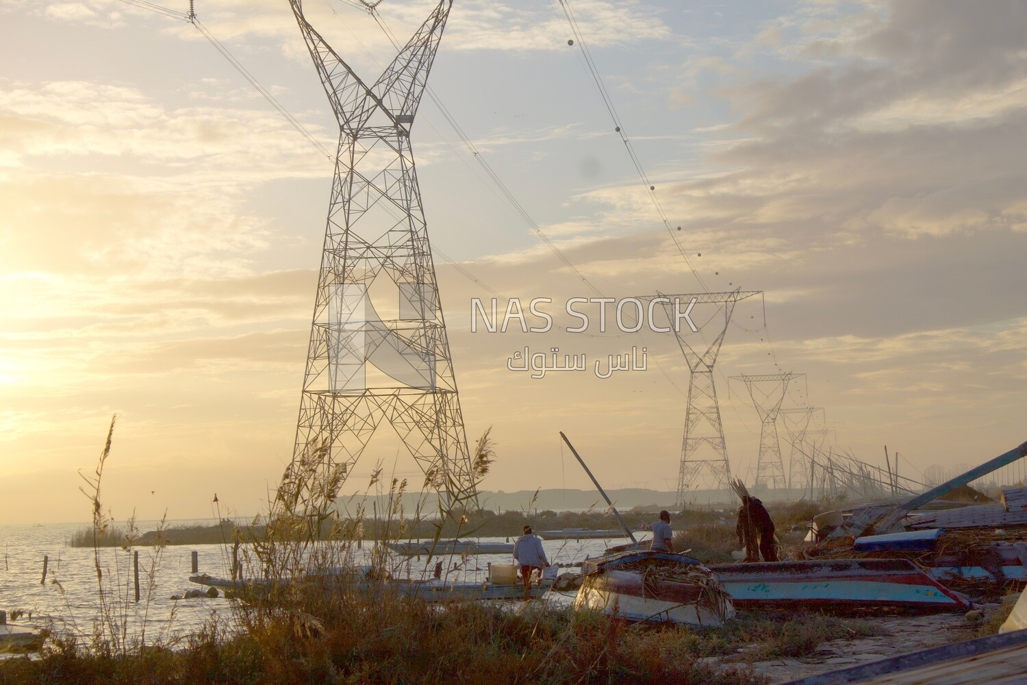 Group of fishermen sit next to their boats in front of the beach and near the high-pressure electric towers during sunset