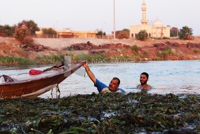 Two men swimming in a river one of them puts his hand on a boat on the beach