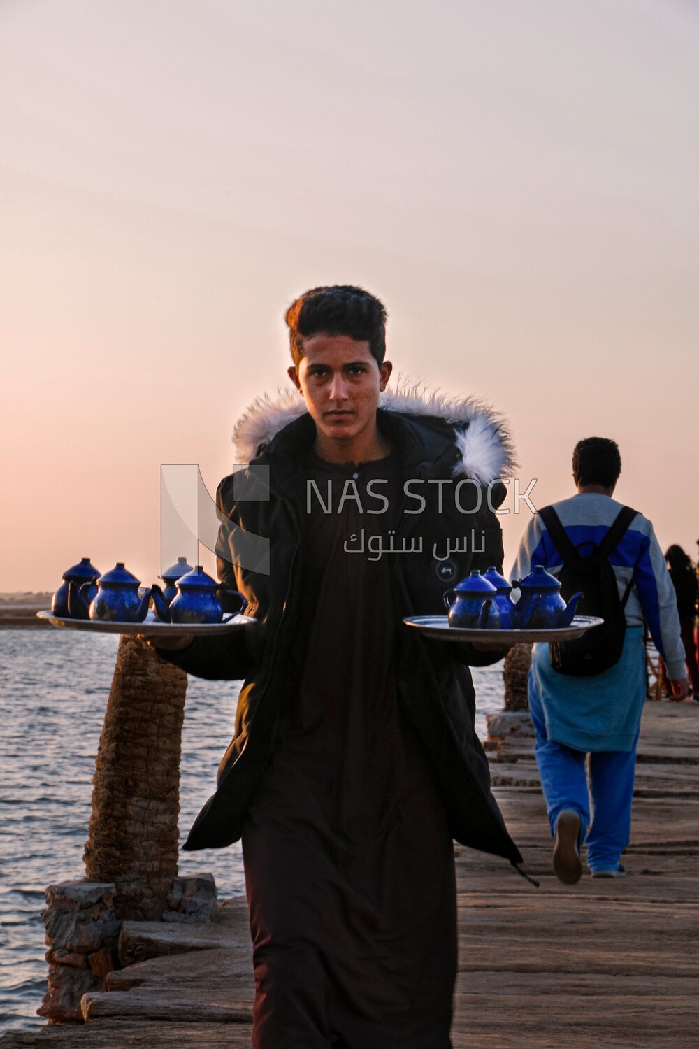 Boy works in a coffee shop holding a tray of tea in front of the sea in Siwa