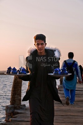 Boy works in a coffee shop holding a tray of tea in front of the sea in Siwa
