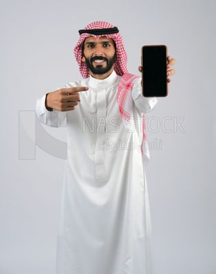 بورتريه مقرب لشاب خليجي مبتسم يرتدي الثوب و الشماغ  يشير باصبعة إلى جهاز الجوال بشاشة سوداء فارغة, برامج التواصل الاجتماعي عبر الهاتف المحمول, مفهوم الاعلانات , استخدام التطبيقات الحديثة و التسويق الألكتروني