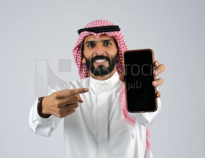 بورتريه مقرب لشاب خليجي مبتسم يرتدي الثوب و الشماغ  يشير باصبعة إلى جهاز الجوال بشاشة سوداء فارغة, برامج التواصل الاجتماعي عبر الهاتف المحمول, مفهوم الاعلانات , استخدام التطبيقات الحديثة و التسويق الألكتروني