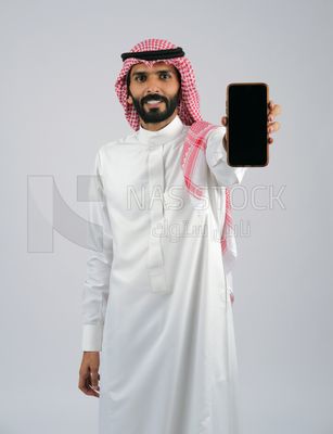 منظر من الامام  لشاب خليجي سعودي مبتسم يرتدي الزى العربى و الشماخ  و يحمل بيده جوال, شاشة سوداء فارغة عبر الهاتف المحمول ,استخدام التكنولوجيا المتطورة، عرض التطبيقات والخدمات الحديثه , جلسات التصوير
