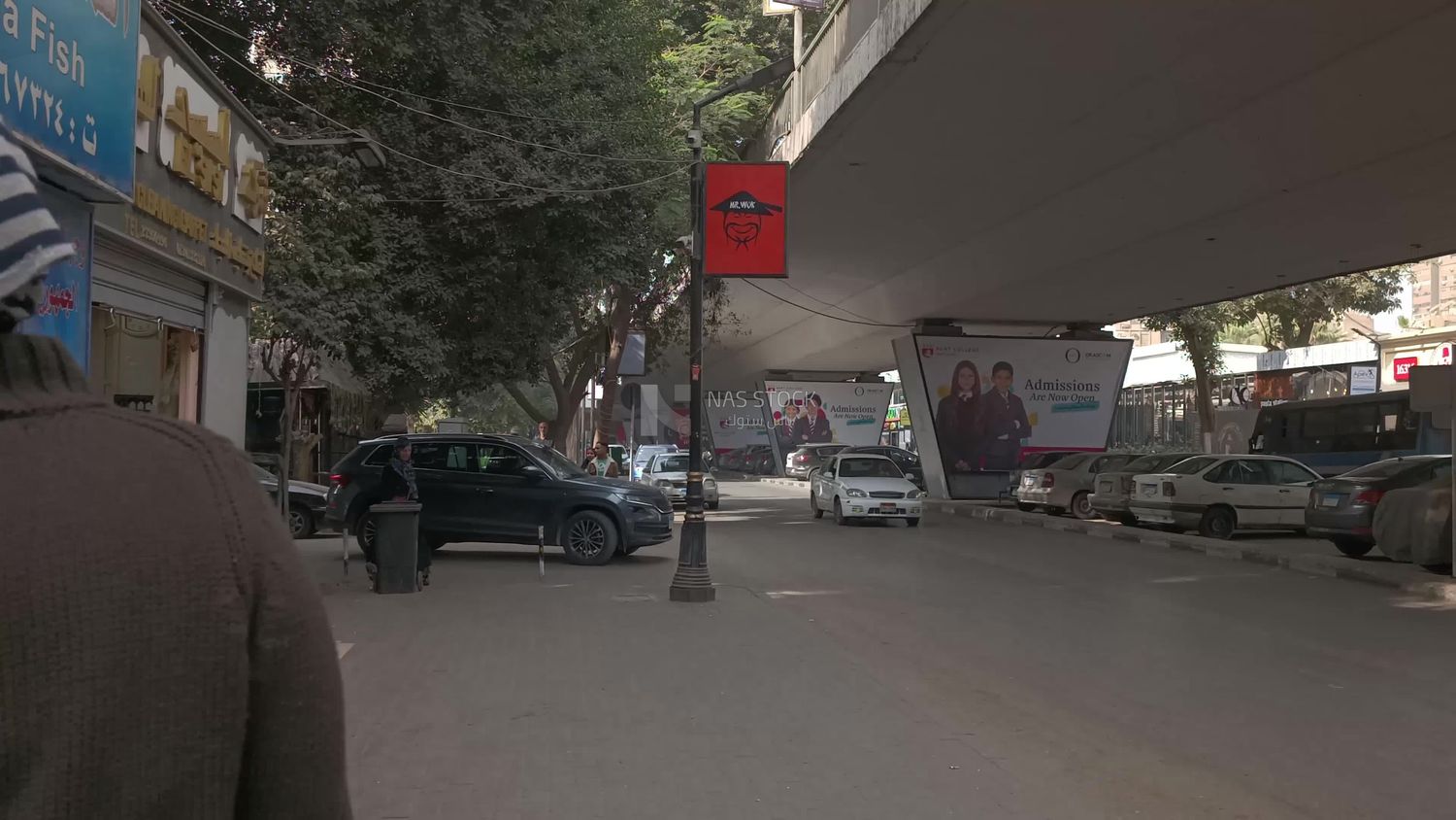 Video showing a street under a bridge where pedestrians and cars pass, and there are shops