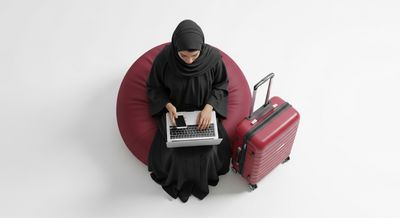 A Gulf girl using a laptop with a red suitcase next to her